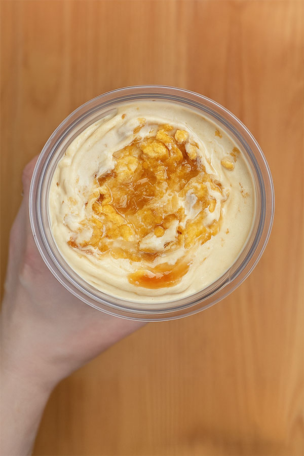 Top-down view of a hand holding a glass bowl of peanut butter caramel crunch protein ice cream, topped with golden caramel sauce and crushed cornflakes