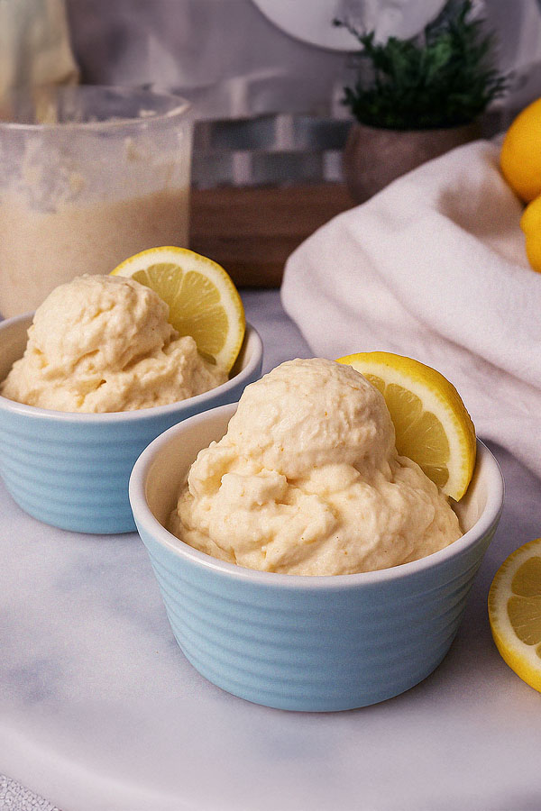 A clear plastic pint filled with lemon cheesecake protein ice cream, topped with a lemon slice. Slices of lemon and whole lemons are placed around it on a textured stone surface