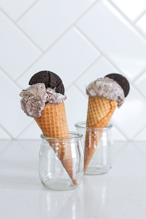 Two cookies and cream ice cream cones standing in glass jars against a white tiled background
