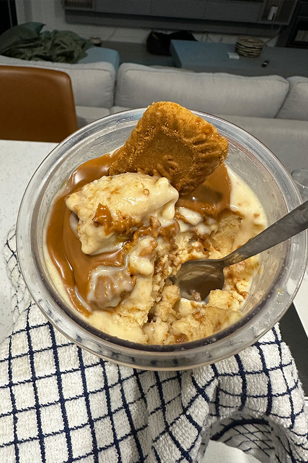 Overhead view of partially eaten Biscoff protein ice cream, topped with melted spread and a biscuit. The container is held over a blue and white checked cloth