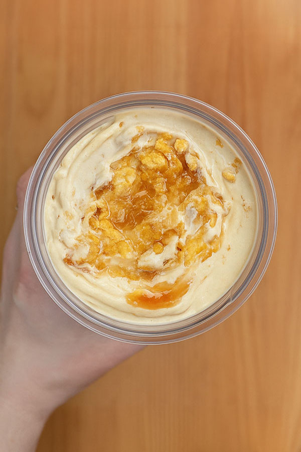 Peanut Butter Caramel Crunch Protein Ice Cream top view Top-down view of a hand holding a glass bowl of peanut butter caramel crunch protein ice cream, topped with golden caramel sauce and crushed cornflakes