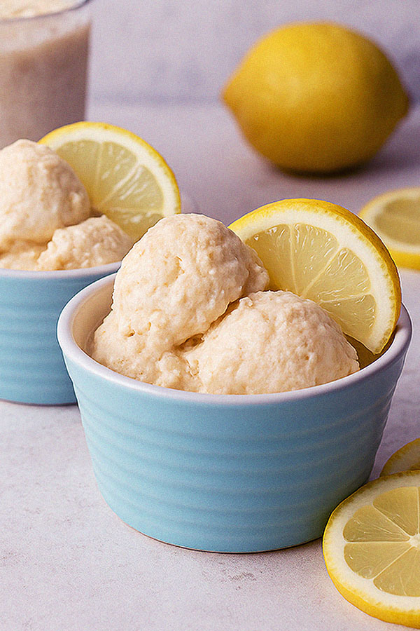 A close-up of two blue bowls with scoops of creamy lemon cheesecake protein ice cream, garnished with lemon slices. A lemon and additional ice cream are softly blurred in the background