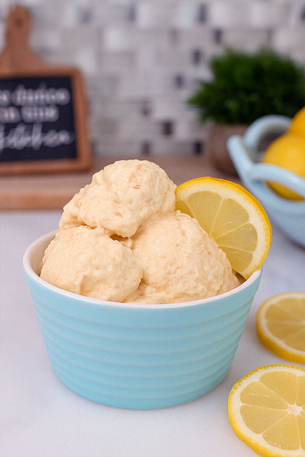 Lemon Cheesecake Protein Ice Cream A single pastel blue bowl filled with smooth lemon cheesecake protein ice cream, topped with a lemon wedge. The background includes a chalkboard kitchen sign, plant, and bowl of lemons