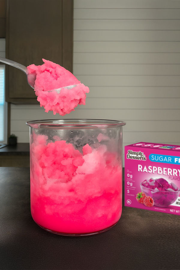 Raspberry Lemonade Sorbet Ninja Creami Pint with pink raspberry lemonade sorbet, with a spoonful lifted above the cup and a Simply Desserts Raspberry Jel mix box in the background on a kitchen counter