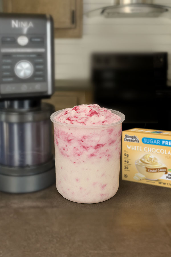 Homemade White Chocolate Raspberry Cheesecake Ice Cream Freshly blended white chocolate raspberry cheesecake ice cream in a Ninja Creami pint container beside a Simply Desserts White Chocolate Pudding Mix box
